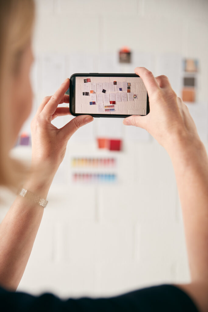 Female Designer Taking Photos Of Designs On Wall With Mobile Phone In Start Up Fashion Business