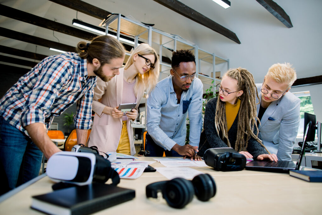 Multiracial business team of young proficient employees leaning on the table together in the modern office room and brainstorming while working on joint project, using tablet and marketing papers.
