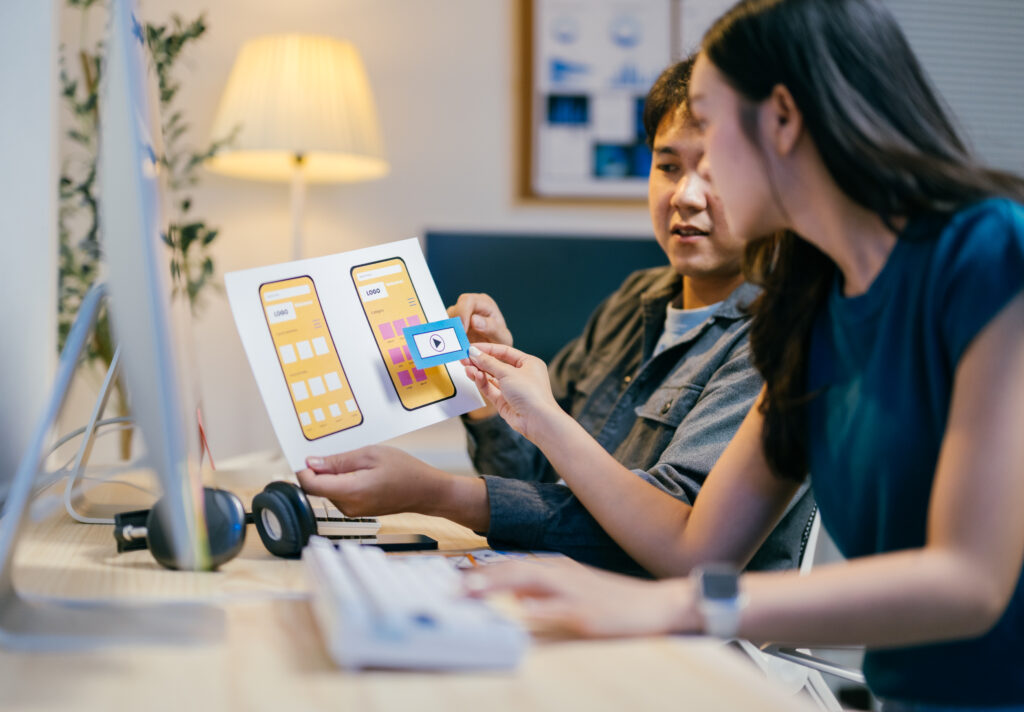 Two asian ui/ux designers are discussing a mobile app wireframe prototype at their desks in the office, improving the user experience with every iteration